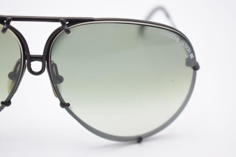 K&ouml;nnte beinhalten: Nahaufnahme einer Vintage-Sonnenbrille von Porsche Design. Die Sonnenbrille hat einen schwarzen Metallrahmen und Verlaufsgl&auml;ser. Der Text "PORSCHE DESIGN" ist auf dem rechten Glas zu sehen. Das Gesamtdesign ist elegant und retro.