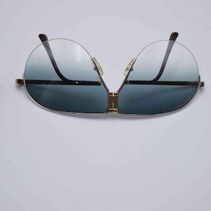 May include: Aviator-style sunglasses with a gold-tone frame and gradient blue lenses. The sunglasses have a double bridge and a sleek, modern design. A burgundy leather case is visible on the left side.