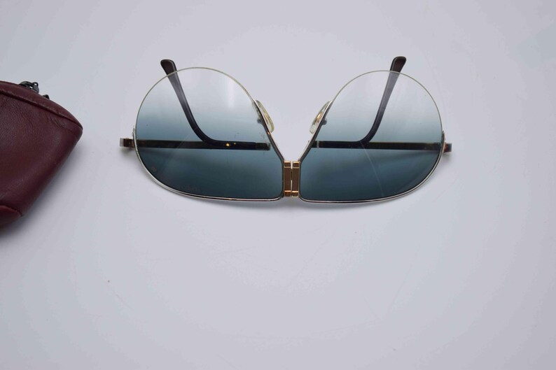 May include: Aviator-style sunglasses with a gold-tone frame and gradient blue lenses. The sunglasses have a double bridge and a sleek, modern design. A burgundy leather case is visible on the left side.