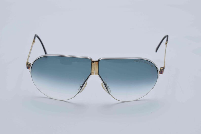 May include: Vintage aviator sunglasses with a gold frame, blue gradient lenses, and black temples. The sunglasses have a classic design with a double bridge and the brand name "Porsche Design" printed on the lens.
