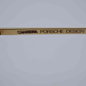May include: Close-up of a gold-toned eyeglass frame. The text "CARRERA PORSCHE DESIGN" is printed in black on the temple arm. The frame has a classic design, with a focus on the brand's logo.
