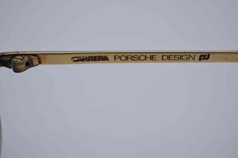 May include: Close-up of a gold-toned eyeglass frame. The text "CARRERA PORSCHE DESIGN" is printed in black on the temple arm. The frame has a classic design, with a focus on the brand's logo.
