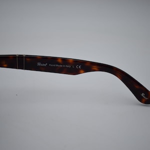 Pode incluir: &Oacute;culos de sol Persol tartaruga com lentes escuras. A haste direita &eacute; vis&iacute;vel, com o logotipo Persol e o texto "Hand Made in Italy L CE". Os &oacute;culos de sol t&ecirc;m uma arma&ccedil;&atilde;o retangular cl&aacute;ssica.