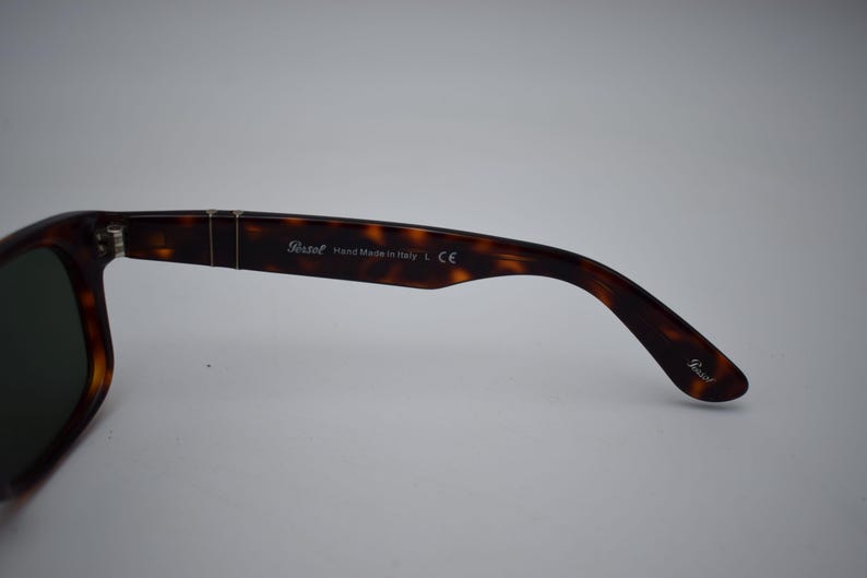 Pode incluir: &Oacute;culos de sol Persol tartaruga com lentes escuras. A haste direita &eacute; vis&iacute;vel, com o logotipo Persol e o texto "Hand Made in Italy L CE". Os &oacute;culos de sol t&ecirc;m uma arma&ccedil;&atilde;o retangular cl&aacute;ssica.