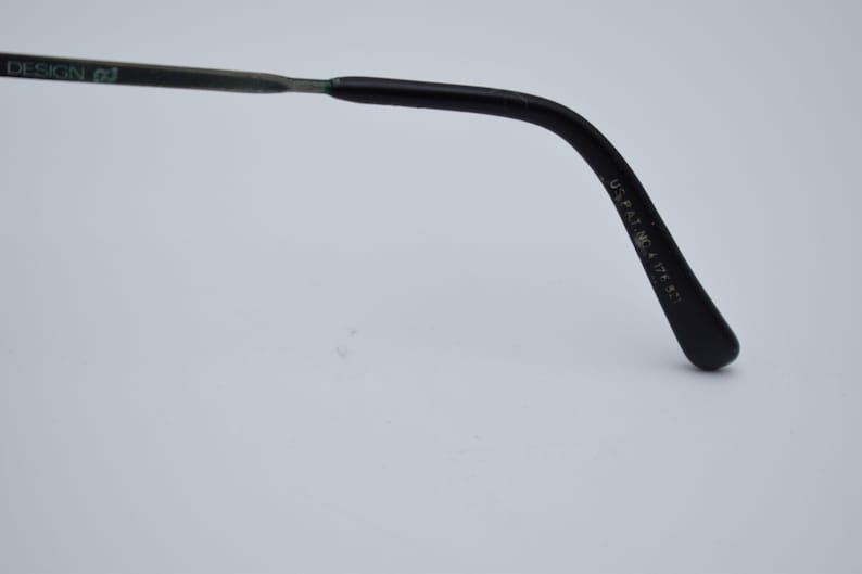 K&ouml;nnte beinhalten: Nahaufnahme des B&uuml;gels einer Brille. Der B&uuml;gel ist schwarz mit einem geschwungenen Design und einer abgerundeten Spitze. Der Text "DESIGN 02" ist auf dem B&uuml;gel sichtbar. Der Text "U.S. PAT. NO. 4,124,871" ist ebenfalls sichtbar.