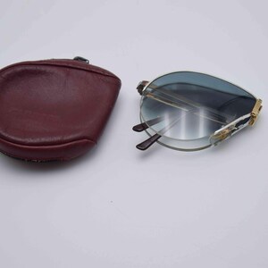 May include: Foldable sunglasses with blue-tinted lenses and a gold-colored frame, next to a burgundy leather case. The sunglasses are partially open, revealing the dark brown arms. The case is oval-shaped.