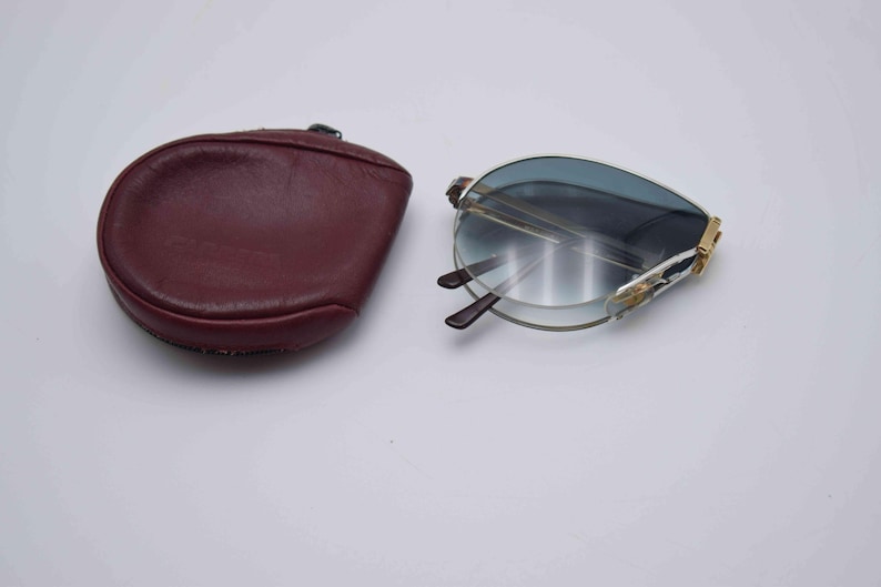 May include: Foldable sunglasses with blue-tinted lenses and a gold-colored frame, next to a burgundy leather case. The sunglasses are partially open, revealing the dark brown arms. The case is oval-shaped.