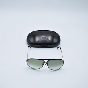 K&ouml;nnte beinhalten: Schwarze Aviator-Sonnenbrille mit gr&uuml;n get&ouml;nten Gl&auml;sern und passendem schwarzem Etui. Die Sonnenbrille hat einen Doppelsteg und d&uuml;nne Metallrahmen. Das Etui ist oval und hat ein geschwungenes Design.