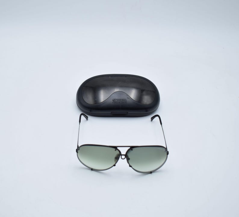K&ouml;nnte beinhalten: Schwarze Aviator-Sonnenbrille mit gr&uuml;n get&ouml;nten Gl&auml;sern und passendem schwarzem Etui. Die Sonnenbrille hat einen Doppelsteg und d&uuml;nne Metallrahmen. Das Etui ist oval und hat ein geschwungenes Design.
