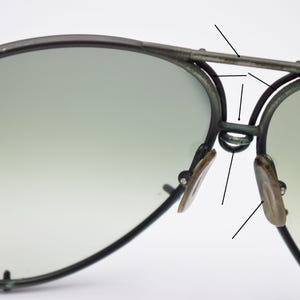 K&ouml;nnte beinhalten: Nahaufnahme einer Pilotenbrille mit dunklem Metallrahmen und Farbverlaufsgl&auml;sern. Die Nasenpads sind hellbraun. Die Sonnenbrille hat ein klassisches Design.