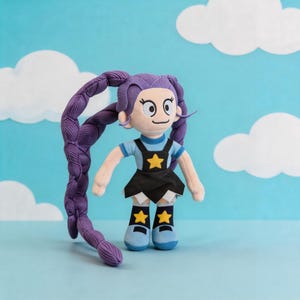 May include: A plush doll with long purple braided hair, wearing a light blue shirt with a yellow star, a black skirt, and matching blue boots with yellow star accents. The doll has large eyes, a simple mouth, and light skin tone fabric.