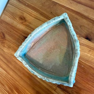 Może przedstawiać: Ręcznie robiony ceramiczny talerz o tr&oacute;jkątnym kształcie i teksturowanej, nieregularnej krawędzi. Wnętrze ma gradient kolor&oacute;w, w tym niebieski, r&oacute;żowy i brązowy. Talerz jest umieszczony na drewnianej powierzchni.
