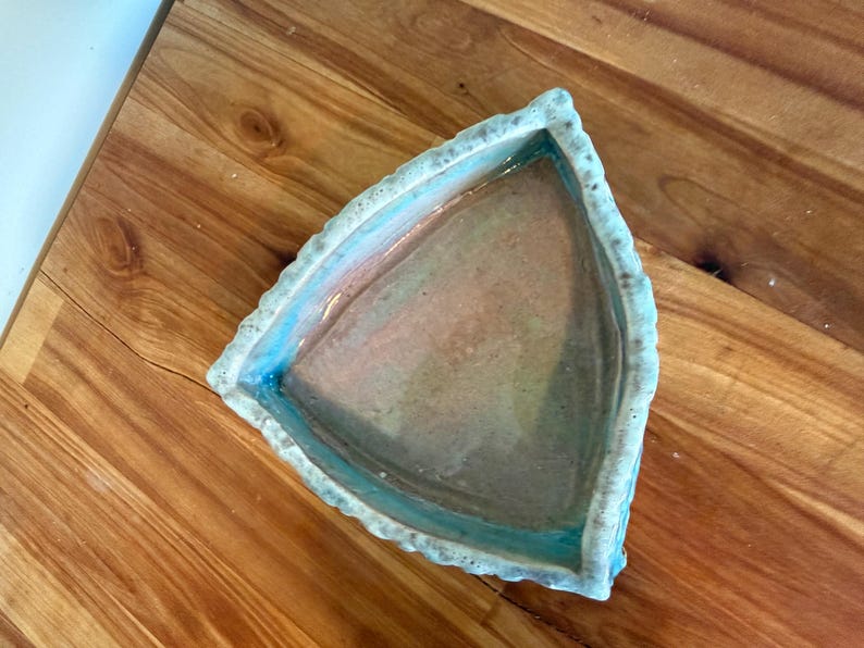 Może przedstawiać: Ręcznie robiony ceramiczny talerz o tr&oacute;jkątnym kształcie i teksturowanej, nieregularnej krawędzi. Wnętrze ma gradient kolor&oacute;w, w tym niebieski, r&oacute;żowy i brązowy. Talerz jest umieszczony na drewnianej powierzchni.