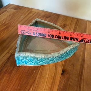Może przedstawiać: Tr&oacute;jkątna ceramiczna miska z turkusowym zewnętrzem i jasnobrązowym wnętrzem. Miska ma około 15 cm długości. Obręcz ma teksturowany wz&oacute;r w kształcie fali. Tekst na linijce brzmi "A SOUND YOU CAN LIVE WITH".