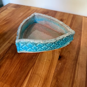 May include: A triangular ceramic planter with a turquoise exterior and a light blue interior. The planter's exterior features a repeating pattern of stylized floral shapes. The interior has a light pink and brown color.
