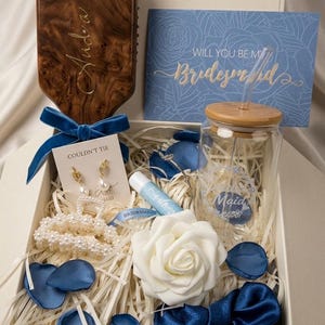May include: A gift box containing a wooden hairbrush, a blue card with the text "Will you be my Bridesmaid", and other items. The box includes a glass with a wooden lid, a white rose, blue petals, and jewellery.