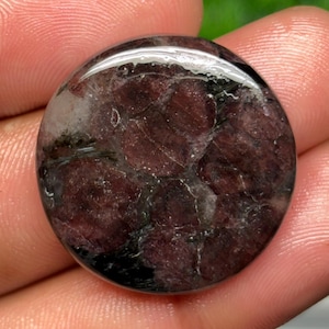Könnte beinhalten: Ein kreisförmiger, polierter Edelstein-Cabochon mit einer tiefroten und kastanienbraunen Farbpalette, durchzogen von komplexen schwarzen Adern und kristallinen Einschlüssen, mit einer glatten, reflektierenden und gewölbten Oberfläche.