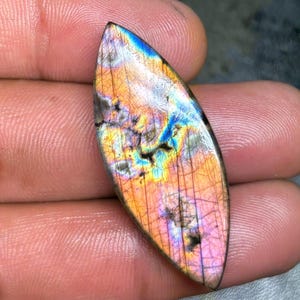 Peut inclure: Une pierre précieuse de labradorite en forme de marquise présentant une surface polie avec des éclats irisés orange, rose, jaune et bleu. La pierre présente des fractures internes naturelles et une silhouette lisse et allongée.