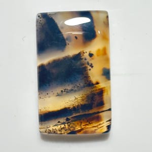 May include: A rectangular polished gemstone cabochon featuring a translucent, creamy beige base with prominent dark blue and black dendritic inclusions that create layered, landscape-like patterns across the smooth, glossy surface of the stone.