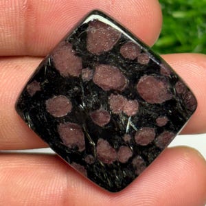 Puede incluir: Una piedra preciosa pulida de forma cuadrada que presenta un color base negro intenso con inclusiones redondeadas de color rojo oscuro incrustadas en toda la superficie. La piedra tiene un acabado liso y brillante y un contraste marcado entre la matriz oscura y las manchas rojas.