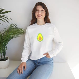 Könnte beinhalten: Weißes Sweatshirt mit einem kleinen, gestickten Avocado-Design auf der Brust. Die Avocado ist grün mit einem gelben Kern und einem lächelnden Gesicht. Das Sweatshirt hat lange Ärmel und einen Rundhalsausschnitt.