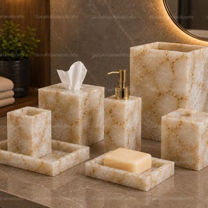 Puede incluir: Un juego de baño de piedra beige con vetas doradas. El juego incluye un dispensador de jabón, una caja de pañuelos, un porta cepillos de dientes, un jabonera y un cubo de basura. Una pastilla de jabón descansa en la jabonera.