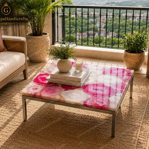 May include: A square coffee table featuring a tabletop made of polished pink and white agate slices with a natural stone pattern. The table is supported by a simple, thin, silver-toned metal frame with four straight legs, creating a clean, modern look.