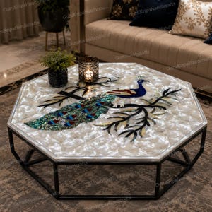 Puede incluir: Una mesa de centro octogonal con una superficie de incrustaciones de nácar blanco y un diseño detallado de pavo real. El pavo real se muestra con plumaje azul, verde e iridiscente, posado sobre ramas negras. Tiene una base de metal negro.