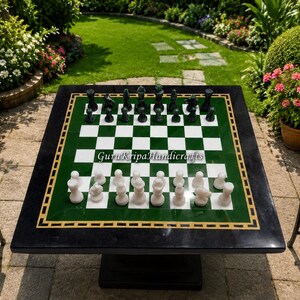 Puede incluir: Una mesa de ajedrez cuadrada de piedra con un tablero a cuadros verde y blanco, un borde negro y una incrustación dorada. La mesa contiene un juego completo de piezas de ajedrez de piedra verde oscuro y blanco. El texto GuruKripaHandicrafts es visible.