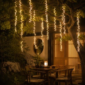 Peut inclure: Un ensemble de guirlandes lumineuses LED blanc chaud suspendues verticalement aux branches d'un arbre, créant un effet de rideau. Les lumières diffusent une lueur douce sur une table et des chaises en bois, avec une lanterne et un service à thé.