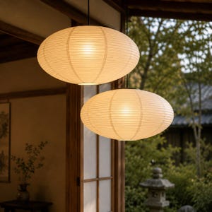 Könnte beinhalten: Zwei cremefarbene, oval geformte Papier-Pendelleuchten hängen von der Decke. Die Lichter sind beleuchtet und werfen einen warmen Schein. Die Lampen hängen in einem Raum mit Holzverkleidung und Blick auf einen Garten.