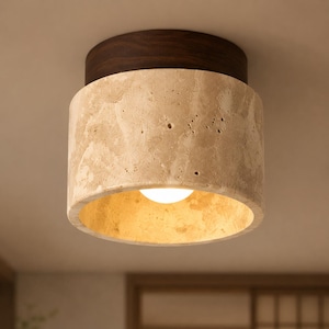 May include: A contemporary ceiling light. It features a cylindrical shade made of textured beige stone, with a dark brown wooden base. The interior of the shade emits a warm light. Designed for flush mounting.