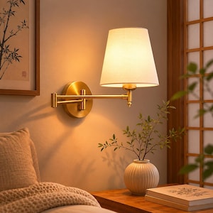 Peut inclure: Une applique murale en laiton avec un abat-jour blanc. La lampe a un bras réglable et une base ronde. Elle est fixée sur un mur beige. Un petit vase avec de la verdure est posé sur une table de chevet en bois.