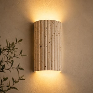 May include: A beige, vertically-oriented wall sconce with a fluted design. The sconce emits a warm, yellow light from the top and bottom, creating a soft glow. The fixture is mounted on a light beige wall, with a hint of greenery in the corner.