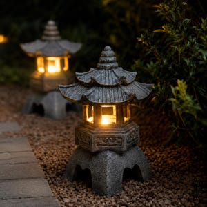 May include: Two grey stone pagoda lanterns with warm interior lighting. The lanterns have a multi-tiered roof and are set on a bed of small stones. The background features green foliage, creating a serene outdoor scene.