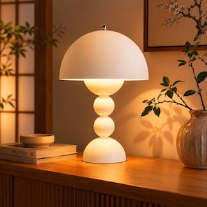 Pode incluir: Uma luminária de mesa branca com uma cúpula arredondada e uma base esférica empilhada. A luminária está iluminada, emitindo um brilho quente. Está sobre uma superfície de madeira, ao lado de um vaso e uma pilha de livros.