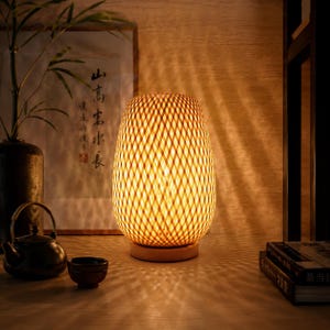 Peut inclure: Lampe de table chaleureuse avec un abat-jour tissé. La lampe, d'environ 30 cm de haut, repose sur un socle en bois, créant des motifs lumineux. La lampe fait partie d'une collection de décoration d'intérieur. Son design évoque la sérénité.