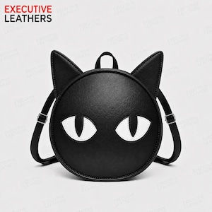 Op de afbeelding: Een ronde zwarte rugzak met twee puntige kattenoren aan de bovenkant en twee witte amandelvormige ogen in het midden. De tas heeft verstelbare schouderbanden met zilverkleurige metalen gespen. De tekst EXECUTIVE LEATHERS is zichtbaar.