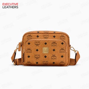 May include: A cognac-colored crossbody bag featuring the MCM monogram print in black. The rectangular bag has a front zippered pocket, a gold-tone metal logo plaque, and a matching adjustable shoulder strap with gold-tone hardware attachments.