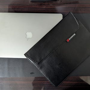 May include: A silver laptop with an Apple logo on the lid positioned next to a black rectangular laptop sleeve featuring a flap closure and a small rectangular tag with the text BLACKSTONE in white letters and a red circular icon.