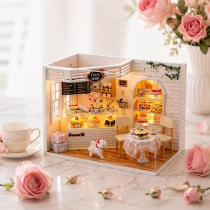 Peut inclure: Une maquette miniature de café avec une façade en briques blanches, exposant des gâteaux et des pâtisseries. Le café comprend une petite table et des chaises, une figurine de chien et une enseigne "CAFE SHOP". Des roses roses et une tasse à thé sont au premier plan.