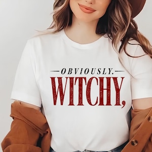 May include: A white short-sleeved t-shirt featuring the black text "OBVIOUSLY." centered above the word "WITCHY," which is printed in a large, bold, dark red font. The shirt has a classic crew neckline and a clean, simple design.