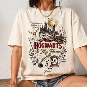 May include: A cream-colored t-shirt featuring a graphic print with the text "HOGWARTS Is My Home", "I solemnly swear that I am up to no good", "9 3/4", "Mischief Managed", and "Always". The design includes a castle, moon, glasses, hat, and books.