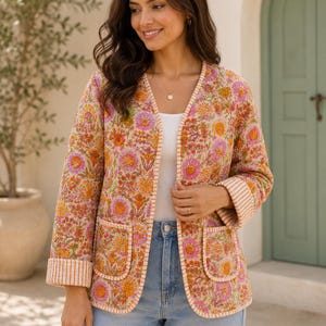Könnte beinhalten: Eine gesteppte Jacke mit einem lebendigen Blumenmuster in Orange-, Rosa- und Gelbtönen. Die Jacke hat eine offene Vorderseite, lange Ärmel mit gestreiften Bündchen und zwei aufgesetzte Vordertaschen. Die Kanten sind mit einer gestreiften Borte versehen.