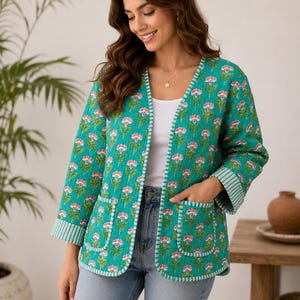 Puede incluir: Una chaqueta acolchada en verde azulado vibrante con un patrón floral repetitivo de flores rosas y blancas. La chaqueta tiene mangas tres cuartos con puños a rayas, frente abierto y dos grandes bolsillos de parche con ribetes a rayas.