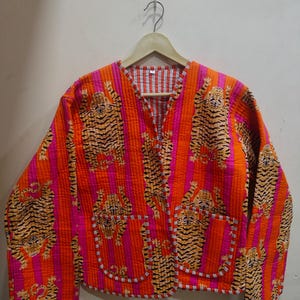 May include: A quilted jacket featuring a vibrant orange and pink vertical striped pattern with a repeating tiger print. The jacket has a V-neckline, long sleeves, and two front patch pockets, all finished with a thin white and black striped trim.