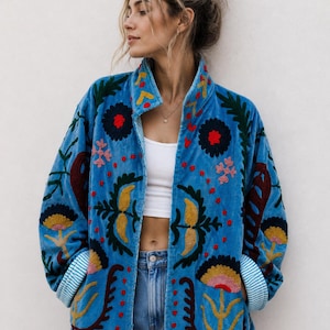 Op de afbeelding: Een felblauw jasje met ingewikkeld, meerkleurig bloemenborduursel in tinten rood, geel en donkergroen. Het jasje heeft een hoge kraag, lange mouwen met lichtblauw en wit gestreepte boorden en een recht, aan de voorzijde open ontwerp.