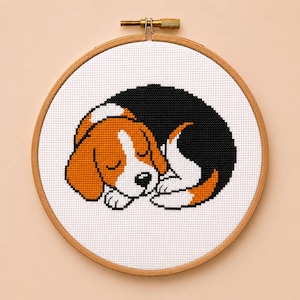 Könnte beinhalten: Ein runder Stickrahmen aus Holz mit einem Kreuzstichmuster eines schlafenden Beagle-Welpen. Der Hund ist mit orangefarbenem, weißem und schwarzem Garn auf weißem Stoff gestickt und liegt zusammengerollt mit geschlossenen Augen da.