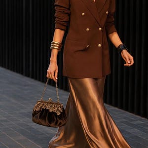 Puede incluir: Un blazer cruzado de color marrón oscuro con botones dorados, una falda larga de satén color bronce metálico y un bolso de terciopelo marrón oscuro con asa de cadena dorada y adornos florales dorados. Se llevan zapatos de tacón con lazos.
