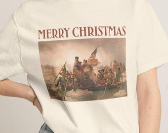 May include: A cream-colored short-sleeve t-shirt featuring a graphic print of the painting Washington Crossing the Delaware. Above the artwork, the text MERRY CHRISTMAS is printed in a dark red serif font. The shirt has a crew neckline.
